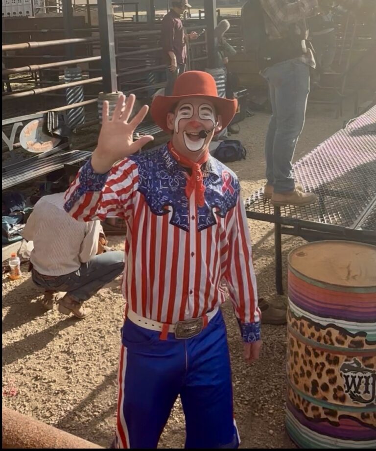 Disco Dalton, a rodeo clown in full face paint and a red cowboy hat, waves to the crowd while wearing a red-white-and-blue striped shirt and blue chaps at a Darby rodeo event, with rodeo pens and a leopard-print barrel in the background.